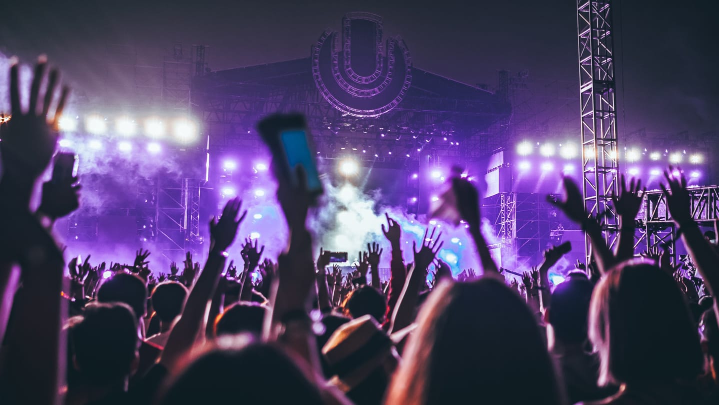 A crowd with hands in the air, gathered in front of an outdoor festival stage at nighttime