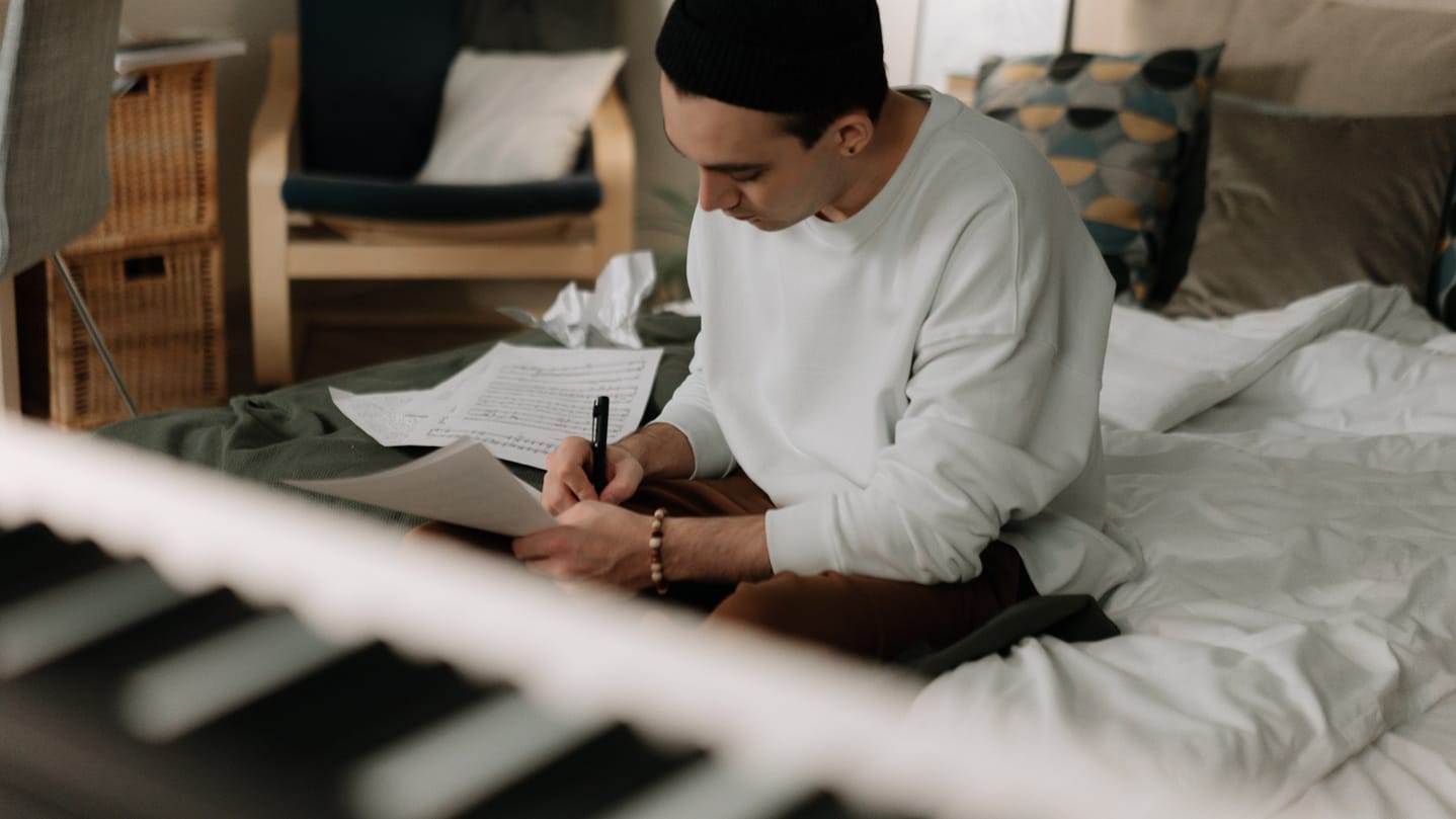 Person sitting on a bed, surrounded by sheets of musical score, writing on a piece of paper