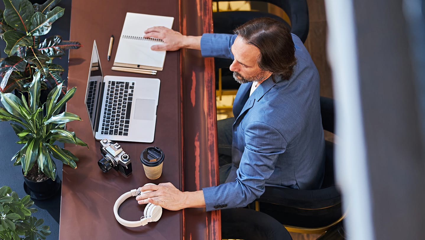Man seated, with notebook, laptop, camera, and headphones laid out on the desk in front of him.