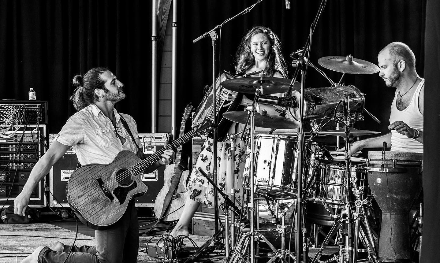 The Adam Ezra band performing on stage, with guitar, drums, and accordion.