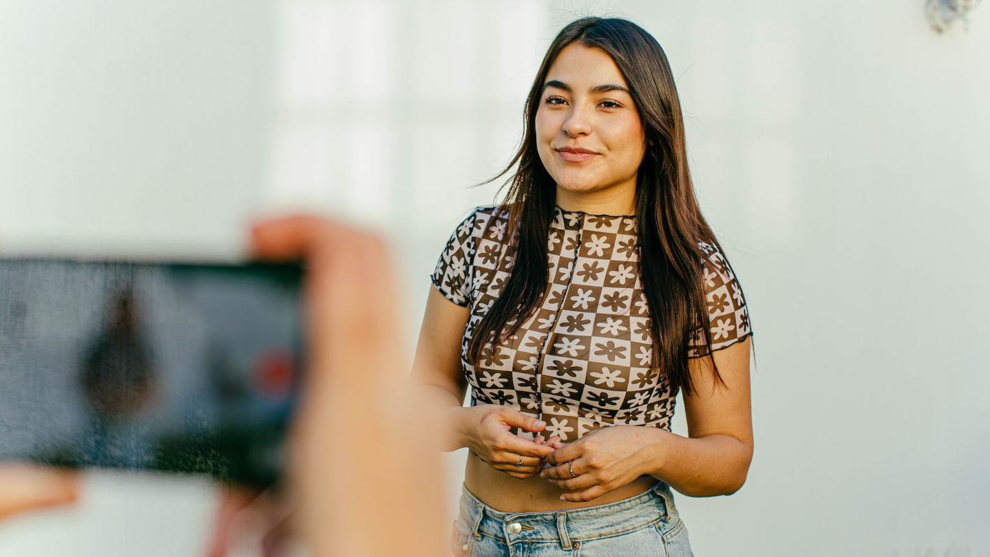 Woman stands against white background; hands are visible holding a smartphone in the foreground to record her.