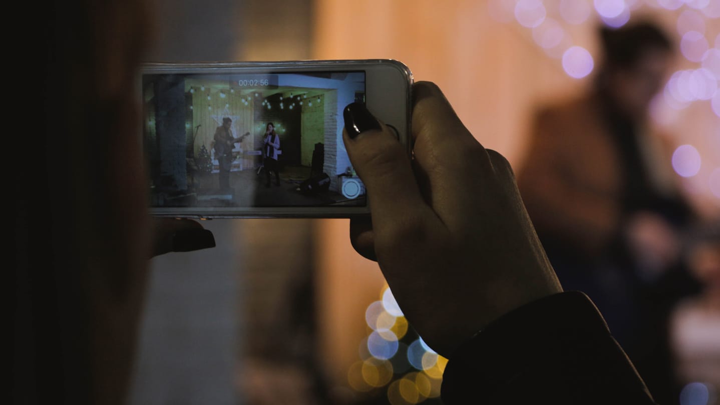 Foreground of smartphone being help up and recording a video of two people performing in a room: guitarist and singer.