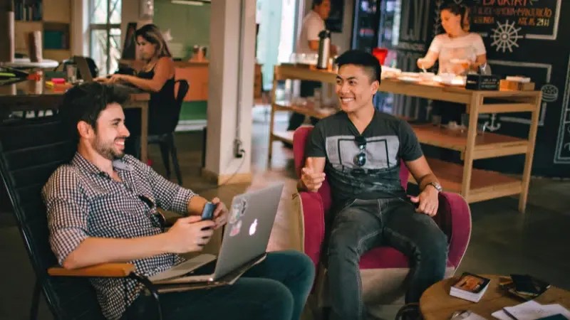 Two men sit and talk in a casual workspace; one holds a laptop while the other gestures, discussing music industry newsletters, with people working in the background.