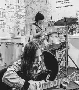 Two young musicians practice in a room filled with posters and notes; one plays electric guitar in the foreground, the other plays drums in the background.