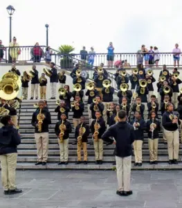 A large school band in black hoodies and khaki pants performs on outdoor steps while people watch from above.