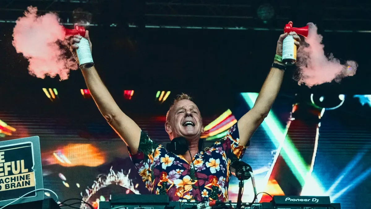 A DJ in a floral shirt, positioned behind a Pioneer DJ setup in front of a colorful screen, enthusiastically raises two pink smoke cannons at a festival. - midnightrebels.com