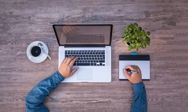 A person working on a laptop with a graphics tablet, coffee, and a plant on a wooden desk, illustrating digital work, creativ
