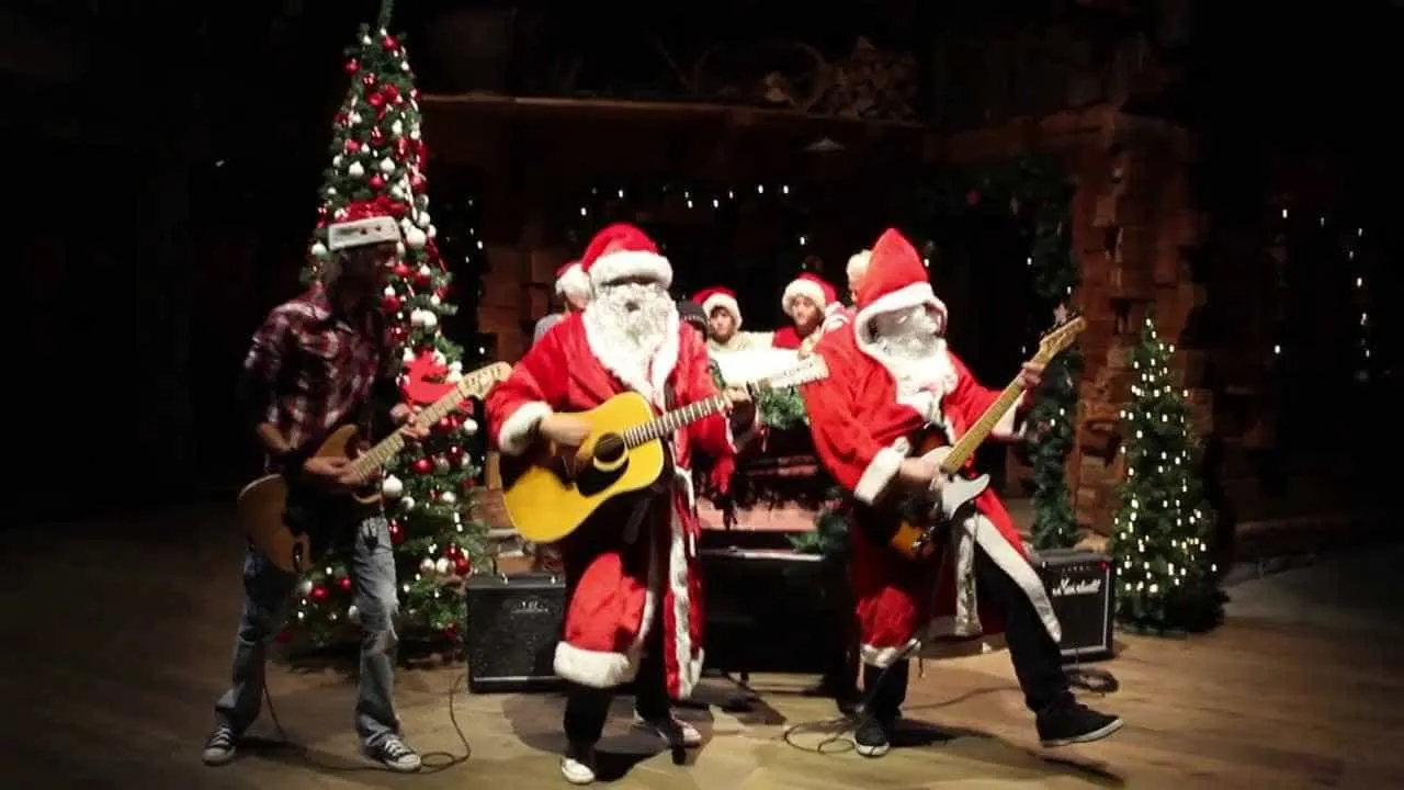 Guitar-playing Santas and musicians performing holiday music on a decorated Christmas stage with trees and festive lights.