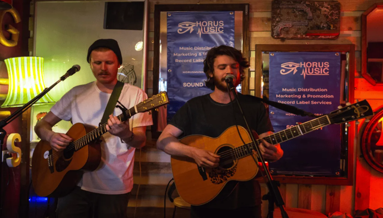 Guitarists performing live at a music venue with "Horus Music" banners, acoustic guitars, and warm ambient lighting.
