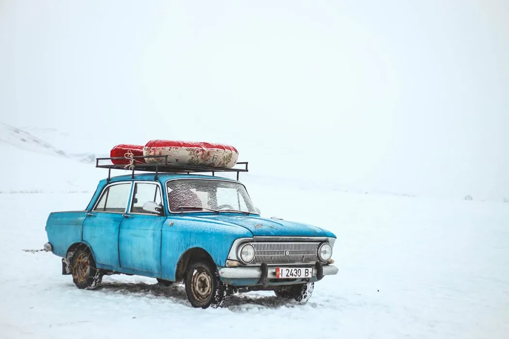 Rusty blue vintage car with a roof rack carrying colorful kayaks, stranded in snow on a vast winter landscape.