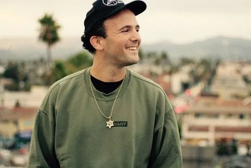 Bright smiling man in casual hoodie and cap outdoors with cityscape background, representing music industry or artist lifesty