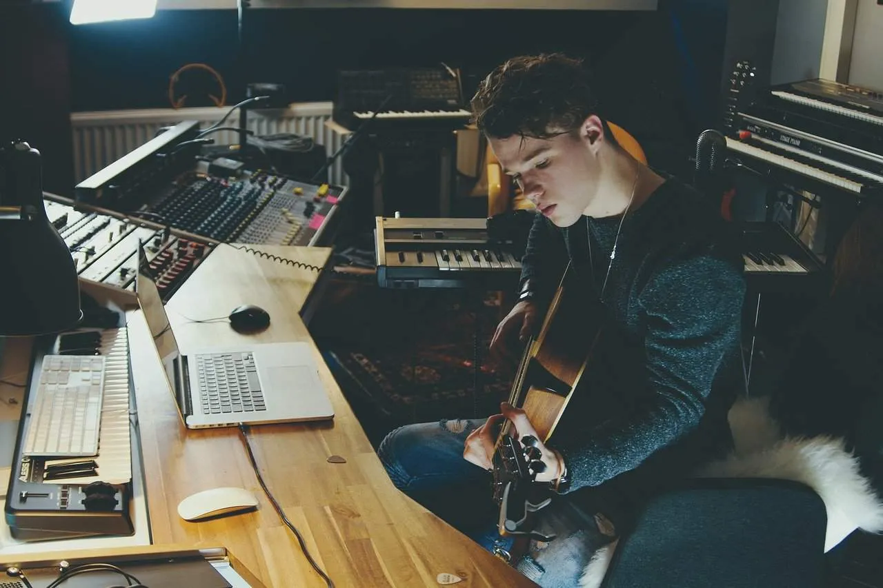 Studio musician working on music production with guitar, laptop, and audio equipment in a modern home recording studio.