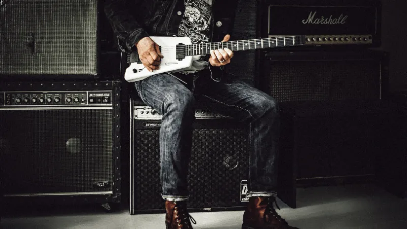 Guitarist playing electric guitar in music studio surrounded by amplifiers and sound equipment.
