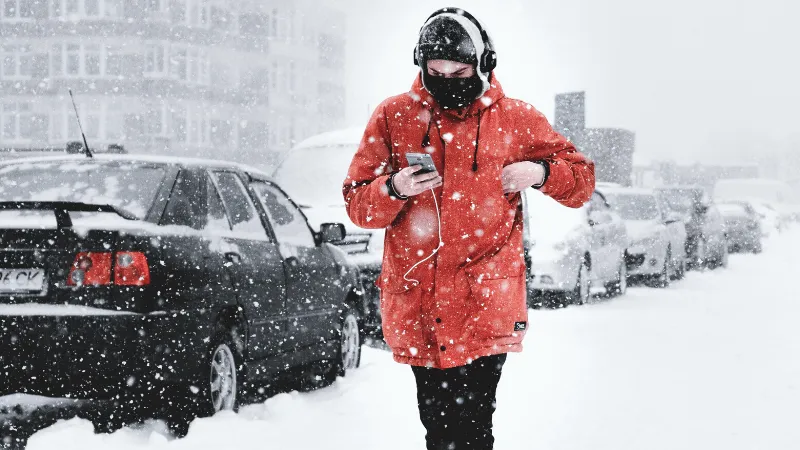 A person in a red winter coat and black face mask walking through a snowy city street while using a smartphone, with parked c