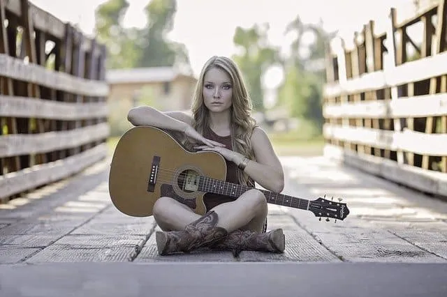 Young woman sitting on wooden bridge holding acoustic guitar outdoors, singer-songwriter, music, casual style, boho vibe, nat