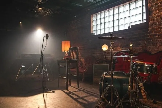 A dimly lit music studio with instruments including a drum set, keyboard, and microphone, featuring exposed brick walls and l