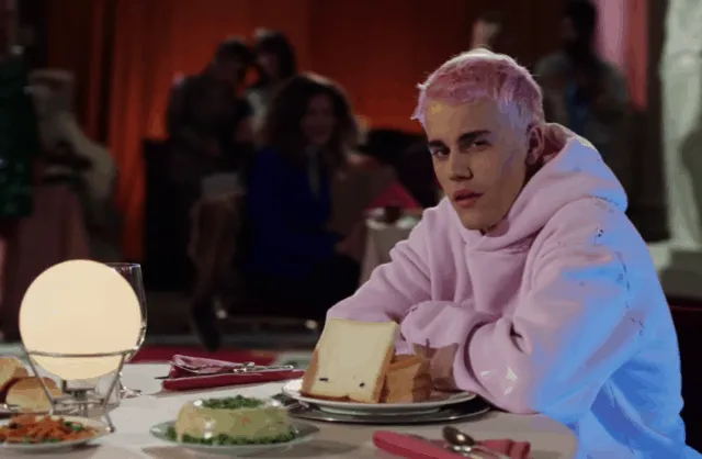 Young man with platinum blonde hair wearing a pink hoodie sitting at a restaurant table with food and drinks during a social