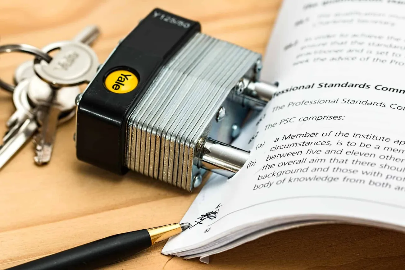 Keys attached to a keychain, a digital lock with a yellow "Vala" sticker, and a printed document with a pen on a wooden surfa
