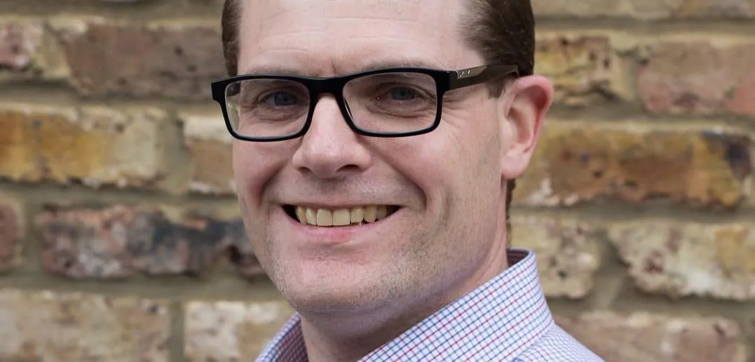 Professional man with glasses smiling in front of a brick wall, representing industry professional, confident speaker, or bus