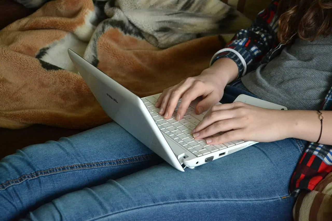 Laptop computer on person's lap, casual indoor setting, young woman typing, cozy environment, tech stay-at-home working or le