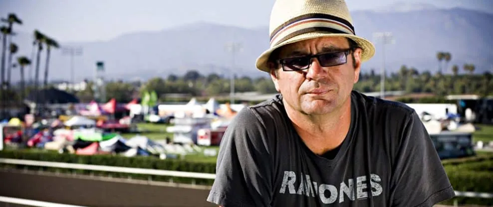 A person wearing sunglasses and a straw hat outdoors at a betting or event venue with tents and palm trees in the background.