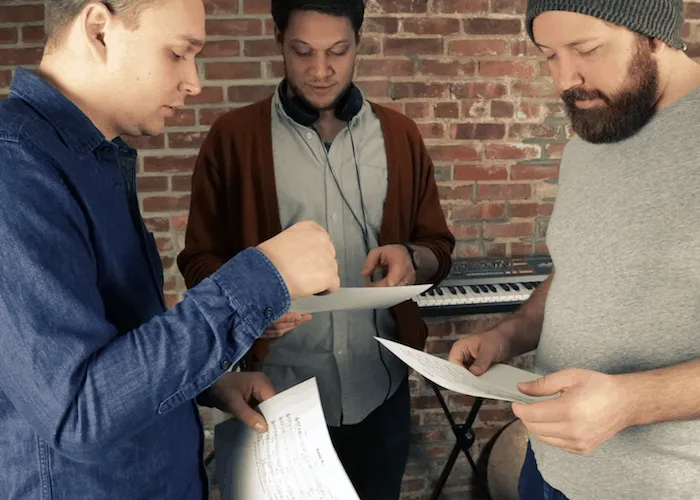 Young music producers collaborating with printed song sheets in a music studio, keyboard in background, brainstorming creativ