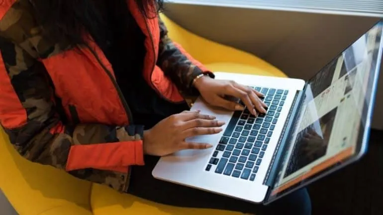 Laptop user typing on keyboard, sitting on yellow couch, working on digital music or social media content, modern workspace,