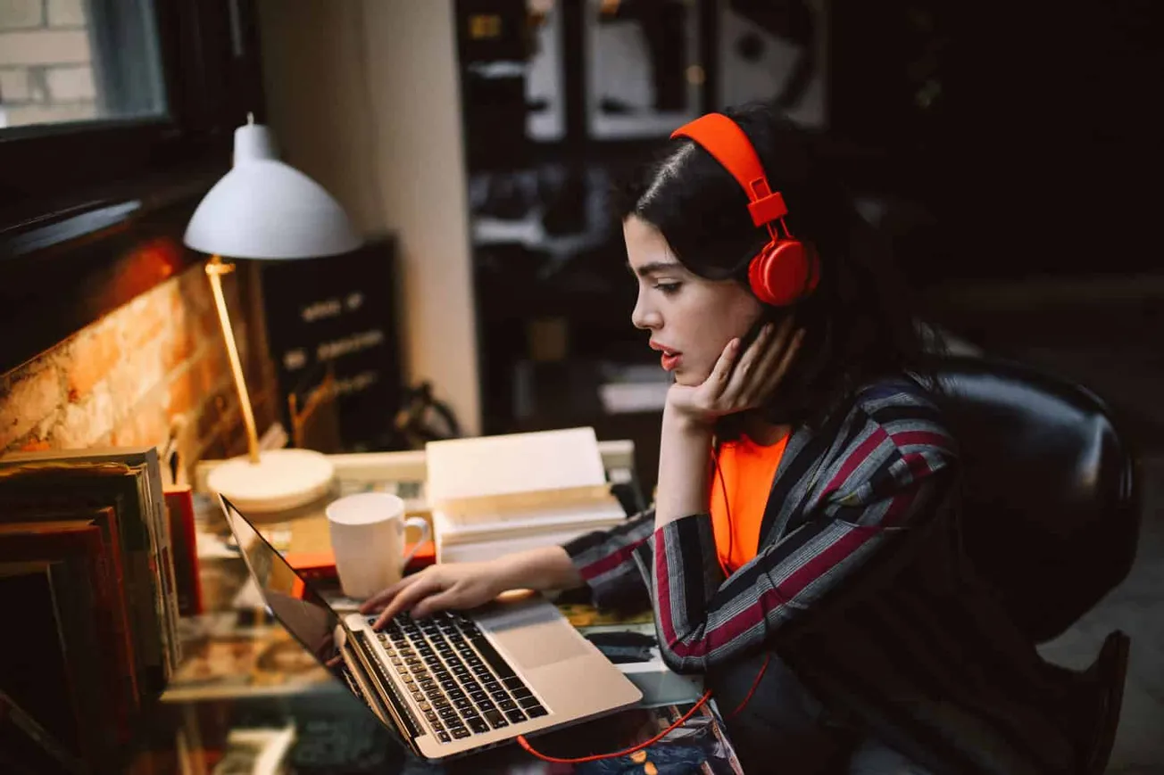 Young woman wearing red headphones working on a laptop at a cluttered desk with books and a coffee mug, in a cozy, dimly lit