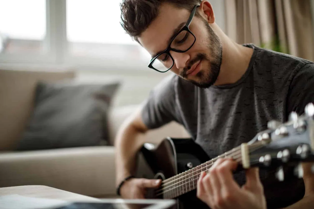 Young man playing acoustic guitar at home, practicing music, relaxing, inner creativity, comfortable living room, musical hob