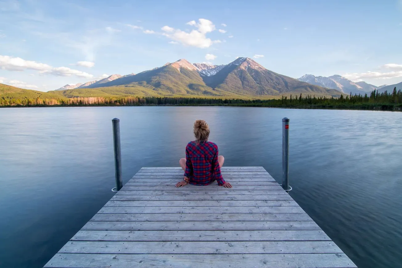 Serene woman sitting on a wooden dock overlooking a tranquil lake with a backdrop of majestic mountains and clear blue sky, c