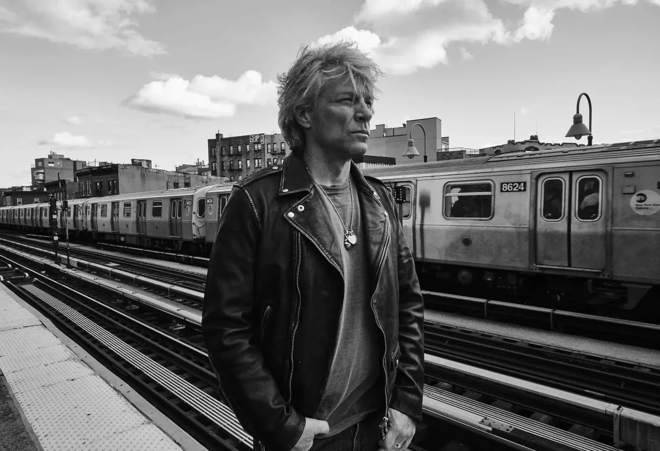 A gritty black-and-white photo of a man with messy hair wearing a leather jacket at a subway station, capturing an urban, edg