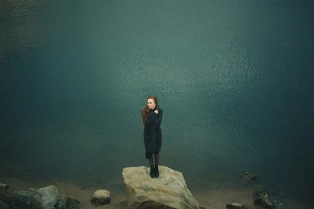 A woman standing on a large rock by a calm body of water, surrounded by smaller rocks, symbolizing reflection and solitude, w