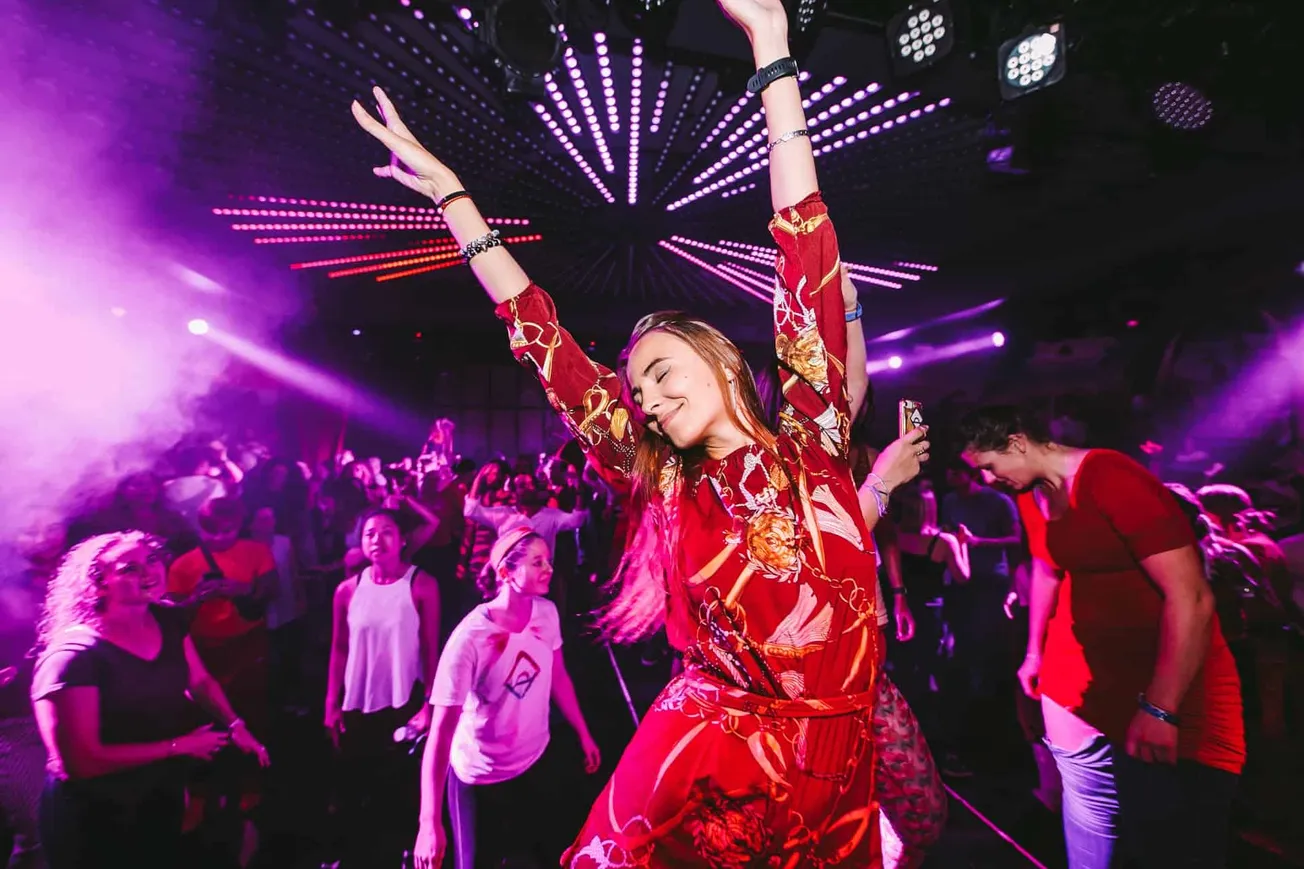 Vibrant woman dancing with eyes closed at a lively nightclub with colorful LED lights and a crowd enjoying music.