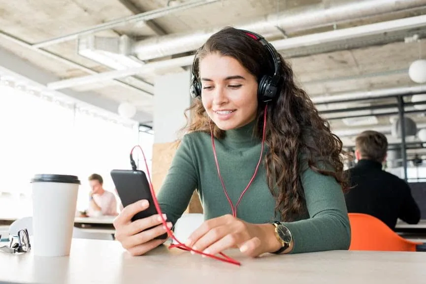 Young woman with headphones using smartphone in a modern coffee shop, enjoying music or a podcast, with a coffee cup on the t