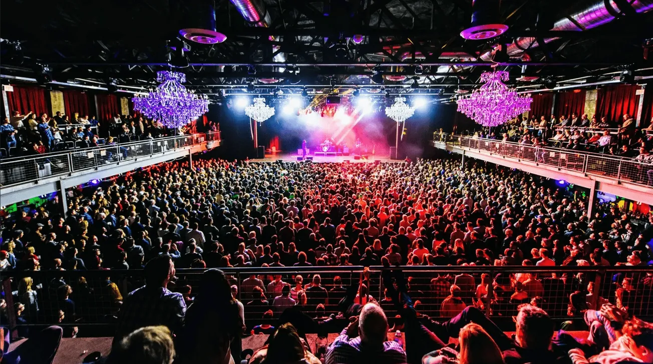 Vibrant concert crowd at an indoor music venue with colorful chandeliers, stage lighting, and an energetic audience enjoying