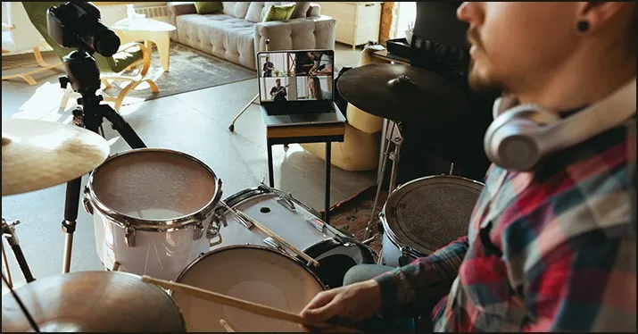 A person practicing drums at home during a virtual band rehearsal, with a laptop displaying remote musicians, set in a cozy l