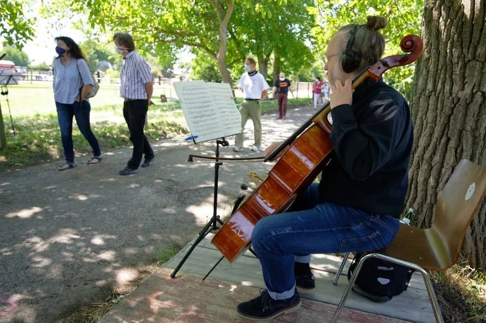 An elderly woman playing a cello outdoors under a large tree, while a small group of people practicing social distancing walk