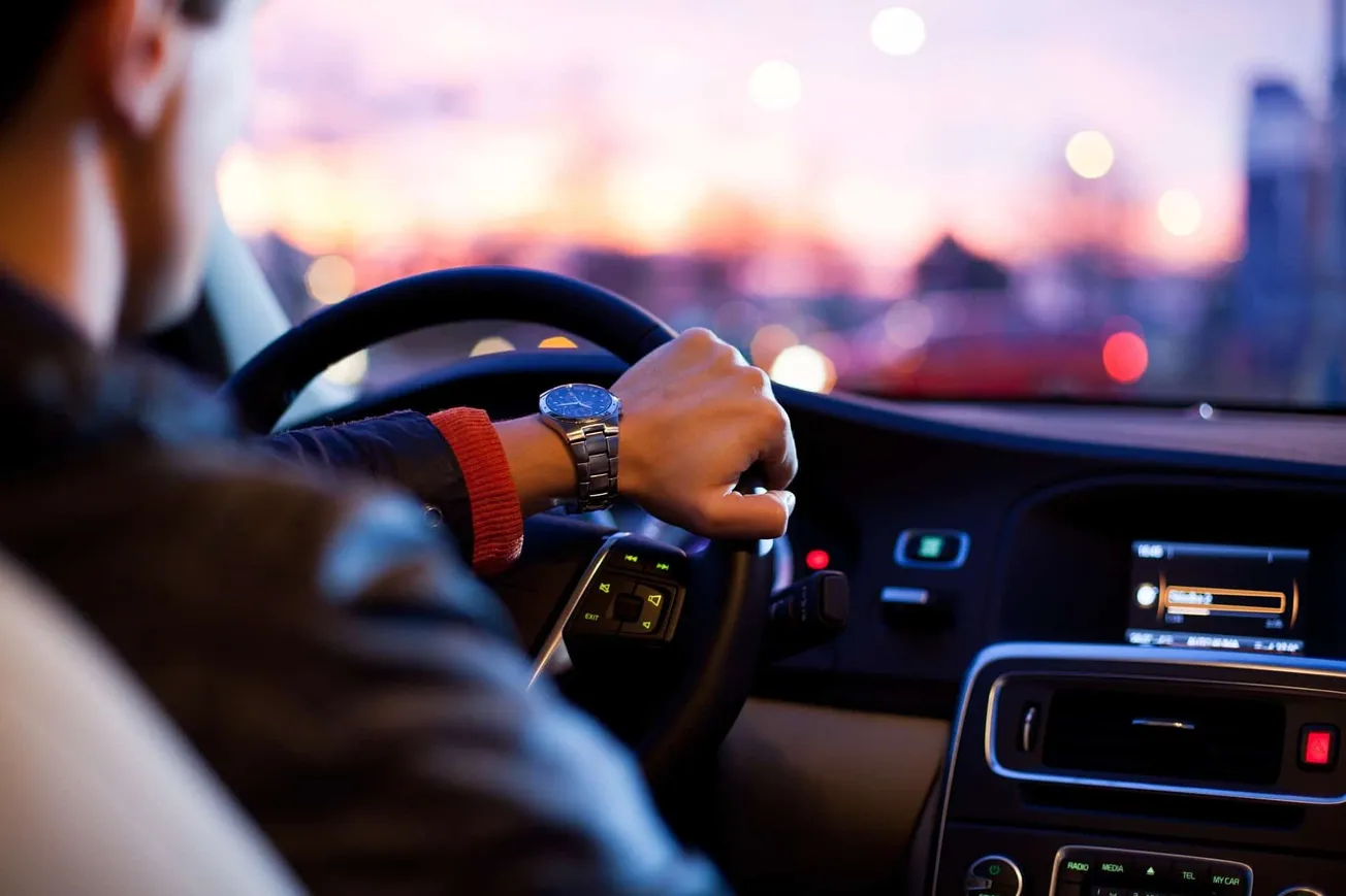 A person driving a car at sunset, showing a close-up of their hand on the steering wheel and wearing a wristwatch, with a blu