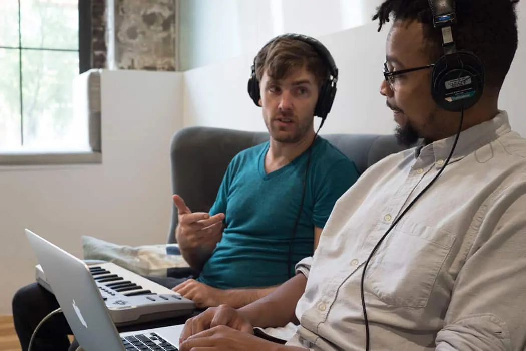 Young men collaborating on music production, using headphones and laptops, in a creative workspace.