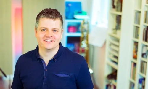 Smiling man in a navy blue shirt standing in a colorful, well-lit room with bookshelves in the background, representing music