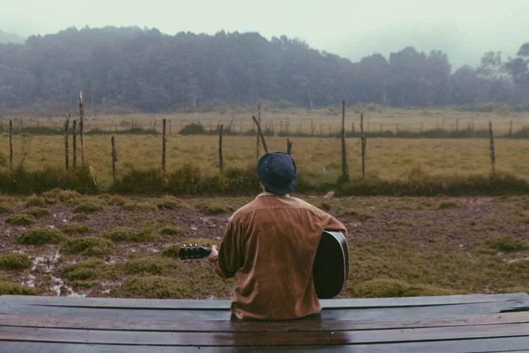 Cityscape, outdoor music performance, person playing acoustic guitar on wooden bench, nature setting with rolling hills, fenc