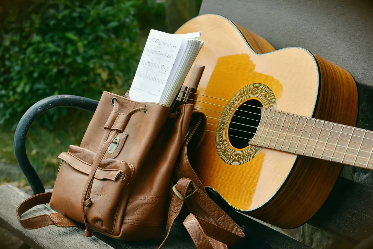A guitar resting against a bench with a brown leather bag and sheet music, outdoors with green foliage in the background.