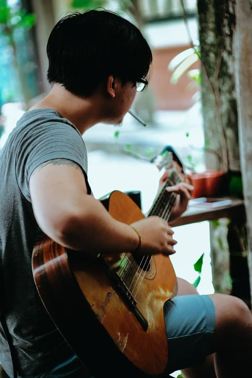 Guitar player playing acoustic guitar, casual musician practicing music, outdoor setting with greenery, young man focused on