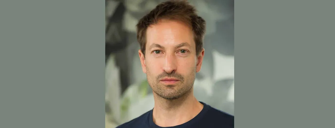 Portrait of a man with brown hair and blue eyes, wearing a black shirt, in an indoor setting with blurred greenery in the bac