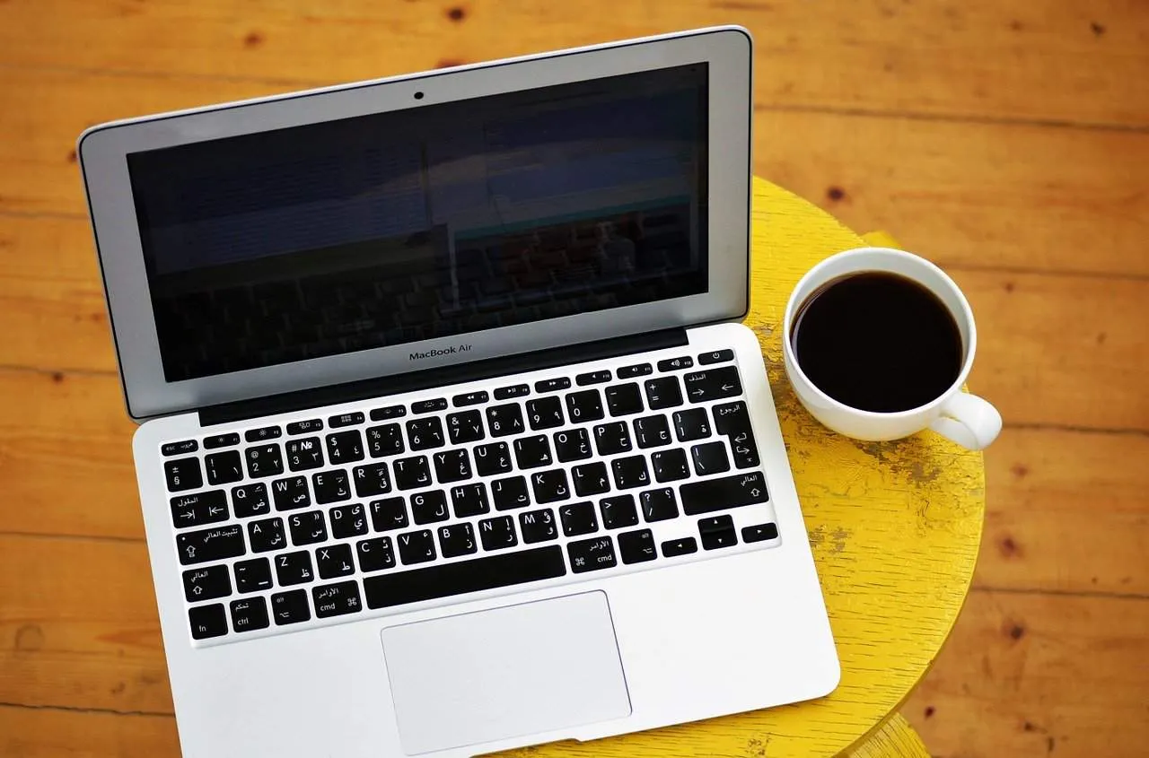 Laptop with Arabic keyboard and cup of coffee on a yellow wooden table, representing digital media, remote work, or tech work
