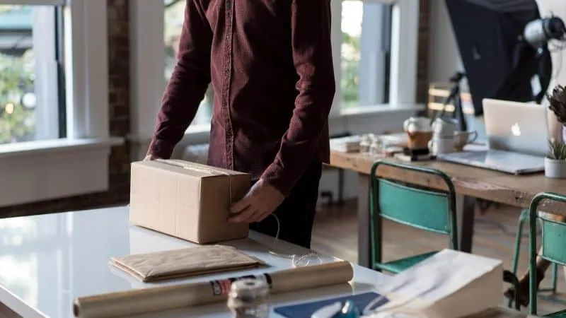 A person preparing a package on a desk in a modern, well-lit office space with large windows, workspace essentials, and a cre