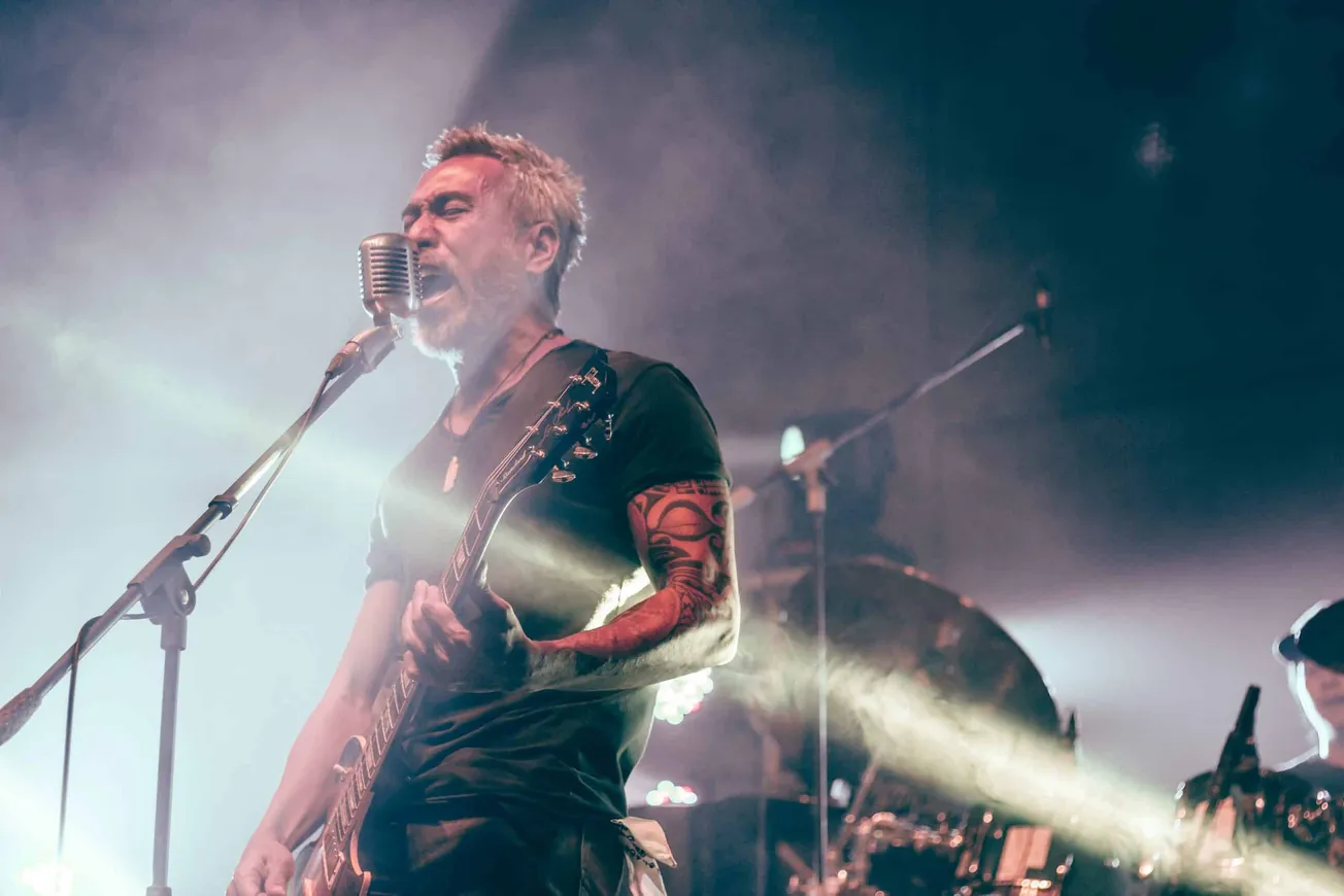 Vocalist guitarist performing live at a music concert, with intense expression and tattoos, illuminated by stage lights, emph