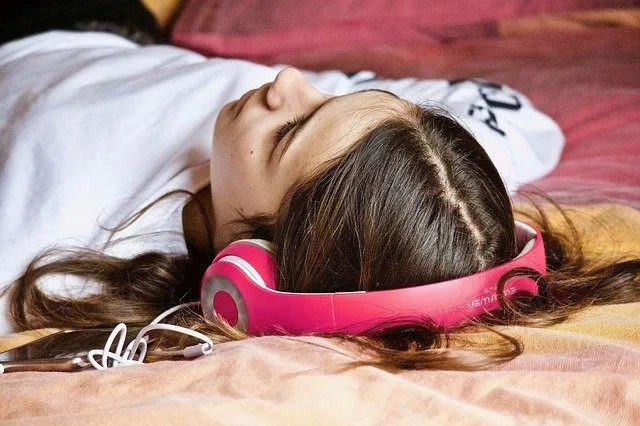 Girl with brown hair lying on bed wearing pink headphones listening to music at home.