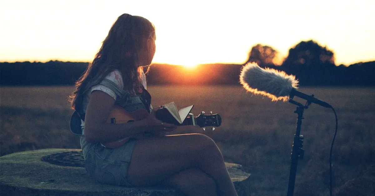 A young woman playing guitar outdoors at sunset, recording vocals with a professional microphone, creating music in a natural