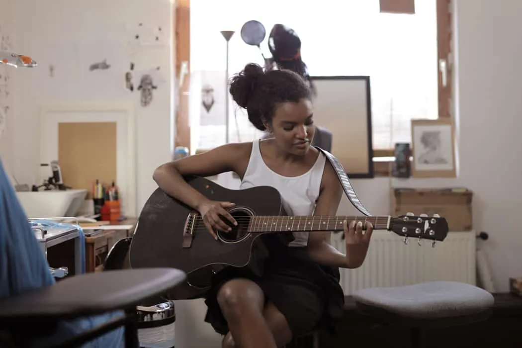 Guitarist woman playing acoustic guitar in cozy music studio bedroom decorated with framed artwork and musical instruments.